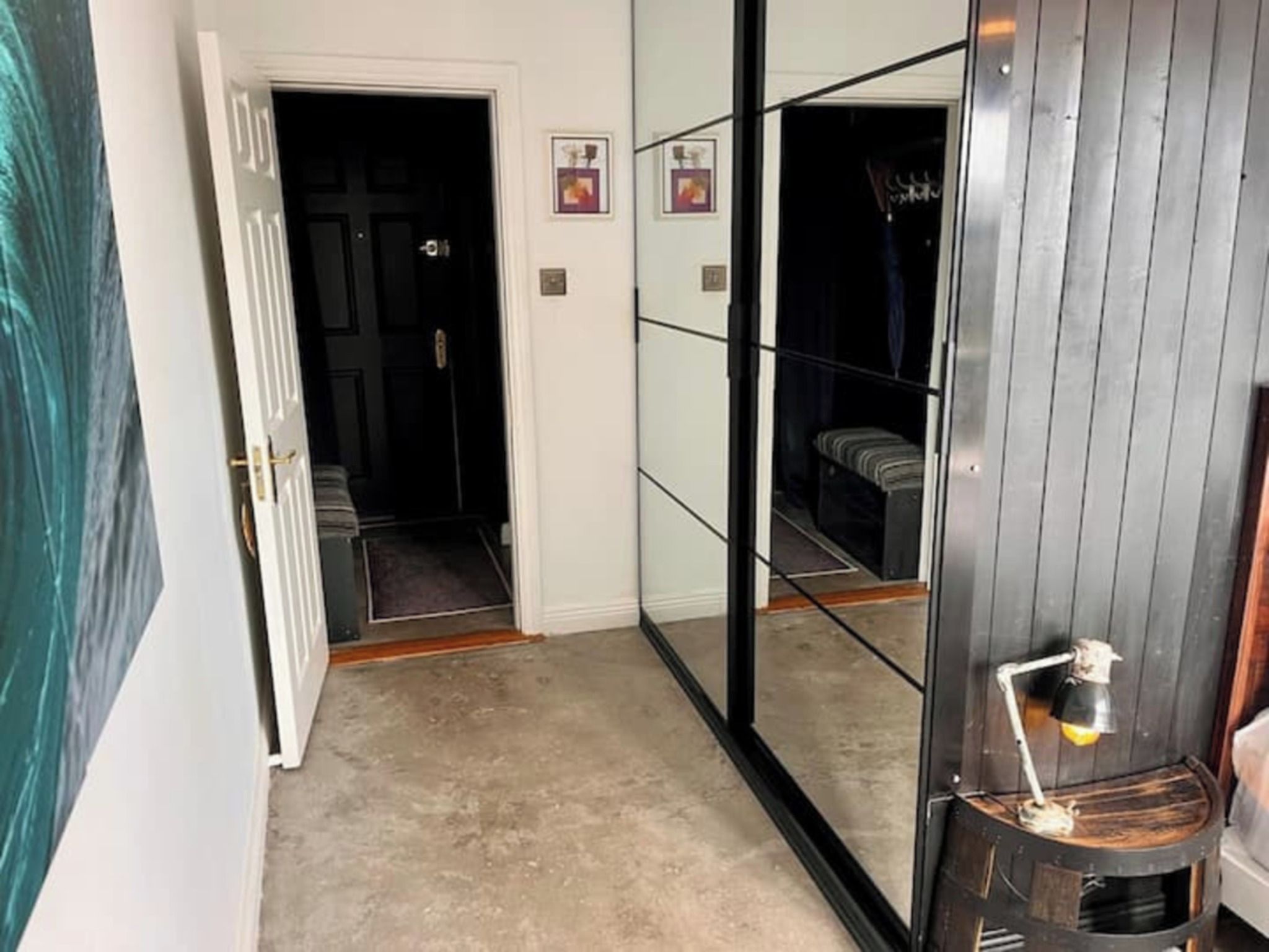 A hallway with a mirrored wall, a door leading to a dark room, and a small table with a lamp.