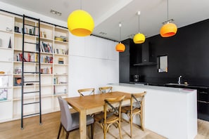 A modern kitchen with a central island, wooden chairs, and a bookshelf filled with books. - Vibrant 2BD Apartment—Parmentier/République (Paris)