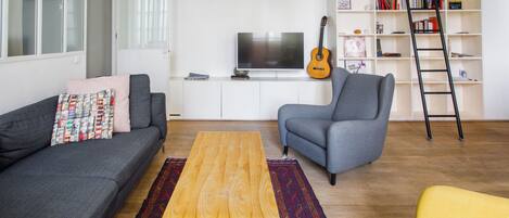 Un salon avec un canapé gris, une table basse en bois, un fauteuil gris, un fauteuil jaune, une bibliothèque, une guitare et un téléviseur.