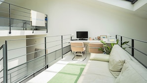 A modern living room with a sofa, a desk, and a television. There is a balcony with a metal railing and a window above the desk. - Stunning 3BD Industrial-style Maisonette—Villette (Paris)