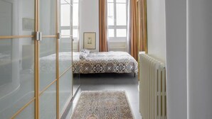 A bedroom with a bed, a radiator, a window with curtains, and a door with glass panels. - Beautiful 1BD Apartment—Porte Dauphine (Paris)