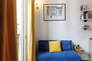 A room with a blue sofa, a yellow pillow, a framed abstract artwork, a bookshelf, and a small side table. - Radiant 1BD Apartment—Clignancourt (Paris)