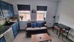 Un espacio habitable compacto con kitchenette, un sofá con cojines, una mesita y una ventana con persianas.