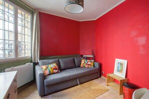A room with a grey sofa, a small wooden table, a framed picture, and a window with decorative iron bars. - Artistic Parisian Home for 5 in Charonne (Paris)