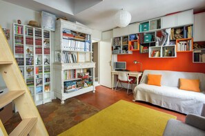 A room with a white desk, a computer, a white sofa with orange cushions, and a bookshelf filled with books and games. - Artistic Parisian Home for 5 in Charonne (Paris)