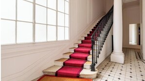 Un magnifique escalier, avec des marches recouvertes de moquette rouge et une balustrade noire, menant à un palier agrémenté d’une colonne blanche.