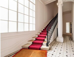 Un magnifique escalier, avec des marches recouvertes de moquette rouge et une balustrade noire, menant à un palier agrémenté d’une colonne blanche.