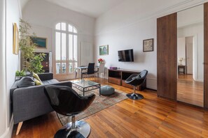 A spacious living room with a large window, a sofa, a coffee table, a TV, and a wooden floor.
