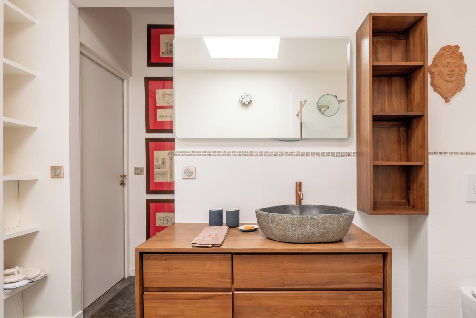 Une salle de bain avec un meuble-lavabo en bois, un grand lavabo en pierre et un miroir au-dessus. Des tableaux encadrés ornent le mur, et une étagère en bois se trouve à droite.