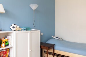 Une chambre avec un lit simple, un bureau avec une lampe et une étagère remplie de jouets et de livres.