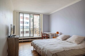 Un dormitorio con cama, escritorio, silla y ventana con vista a edificios.