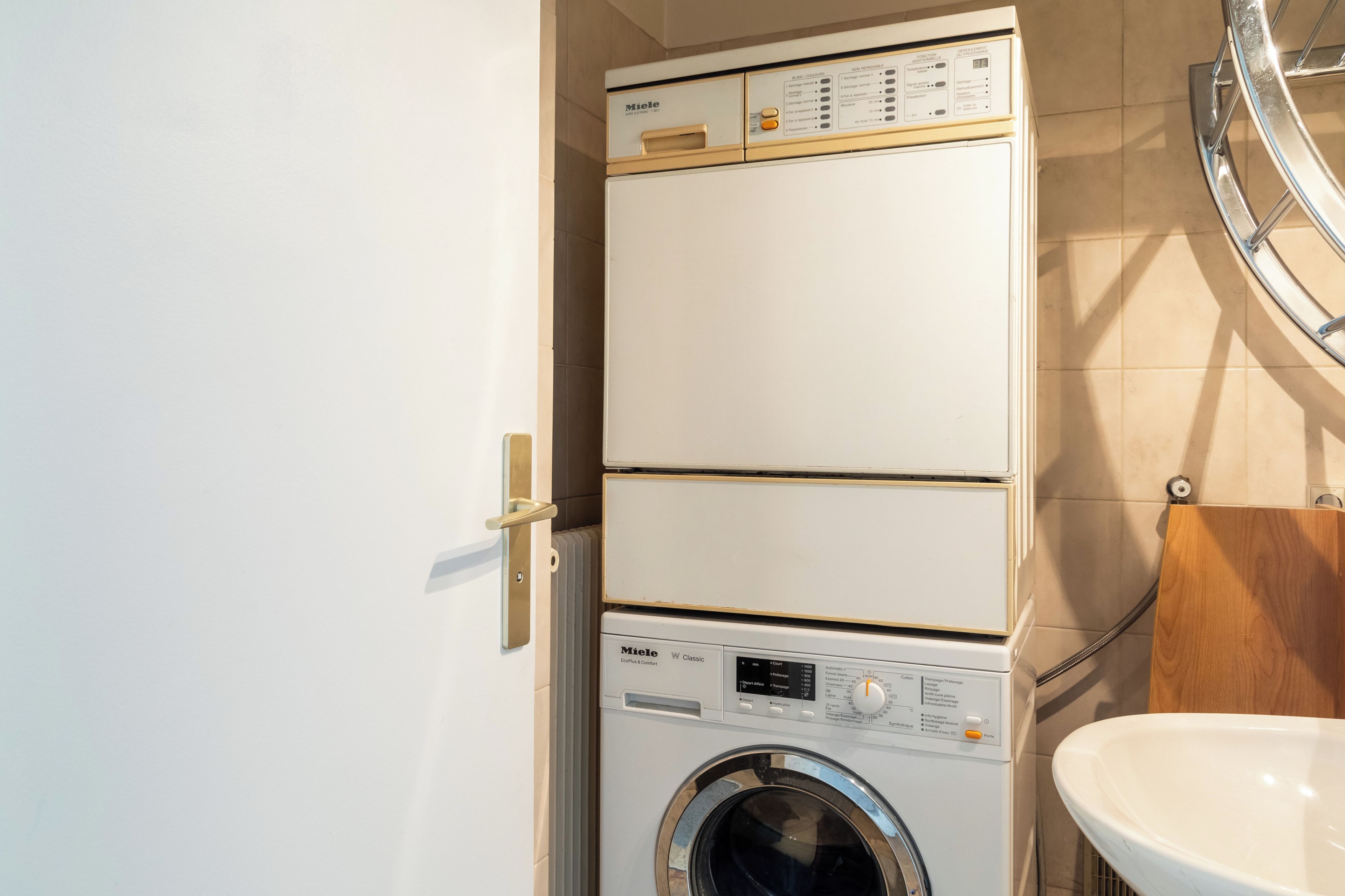Un lave-linge compact, blanc, à deux portes, doté d’un panneau de commande et d’une porte à chargement frontal, installé dans une salle de bain avec un lavabo blanc et un meuble en bois.