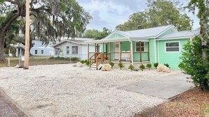 A green house with a porch, surrounded by gravel and trees.