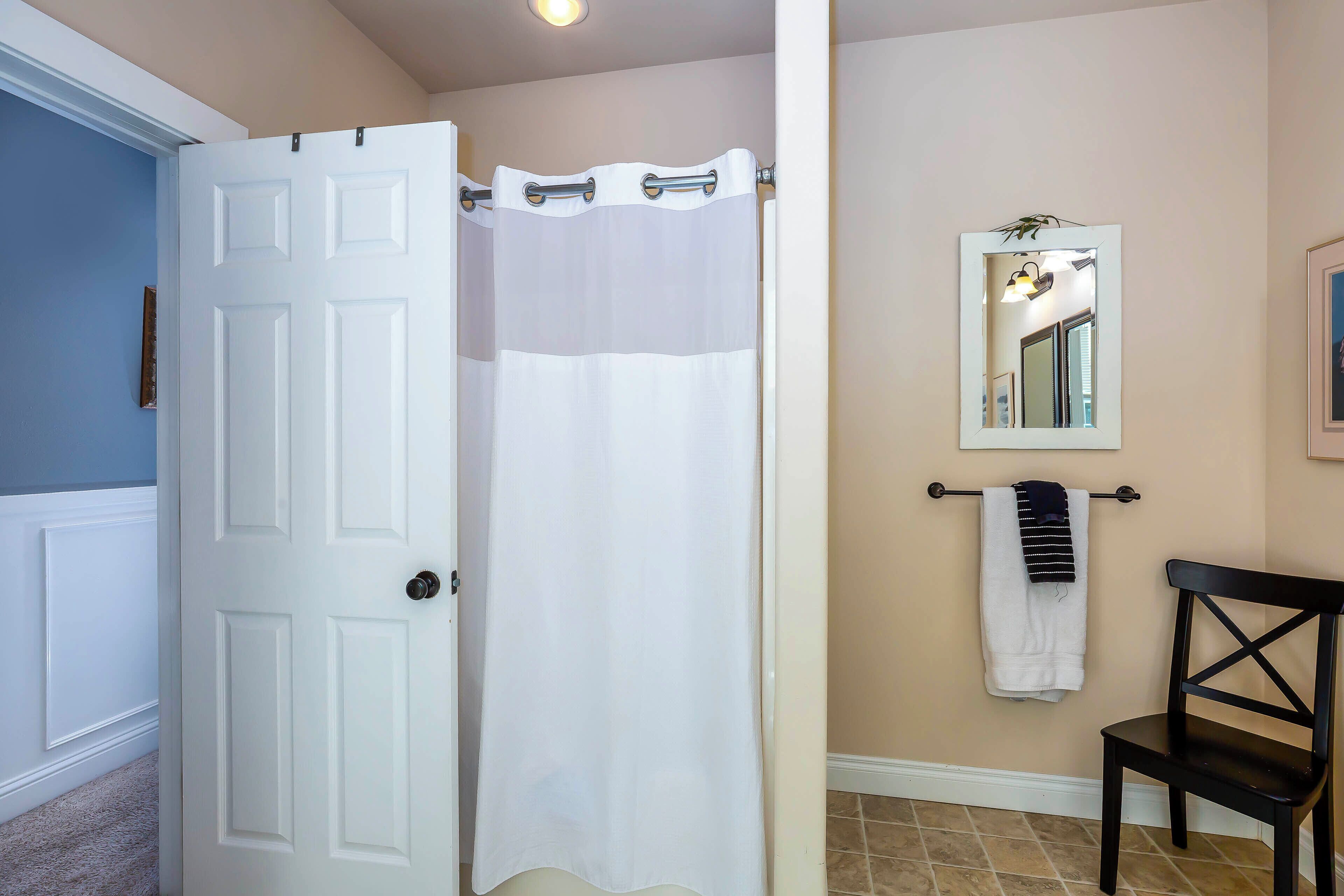 A bathroom with a white door, a shower curtain, a towel rack with a towel, a mirror, and a chair.