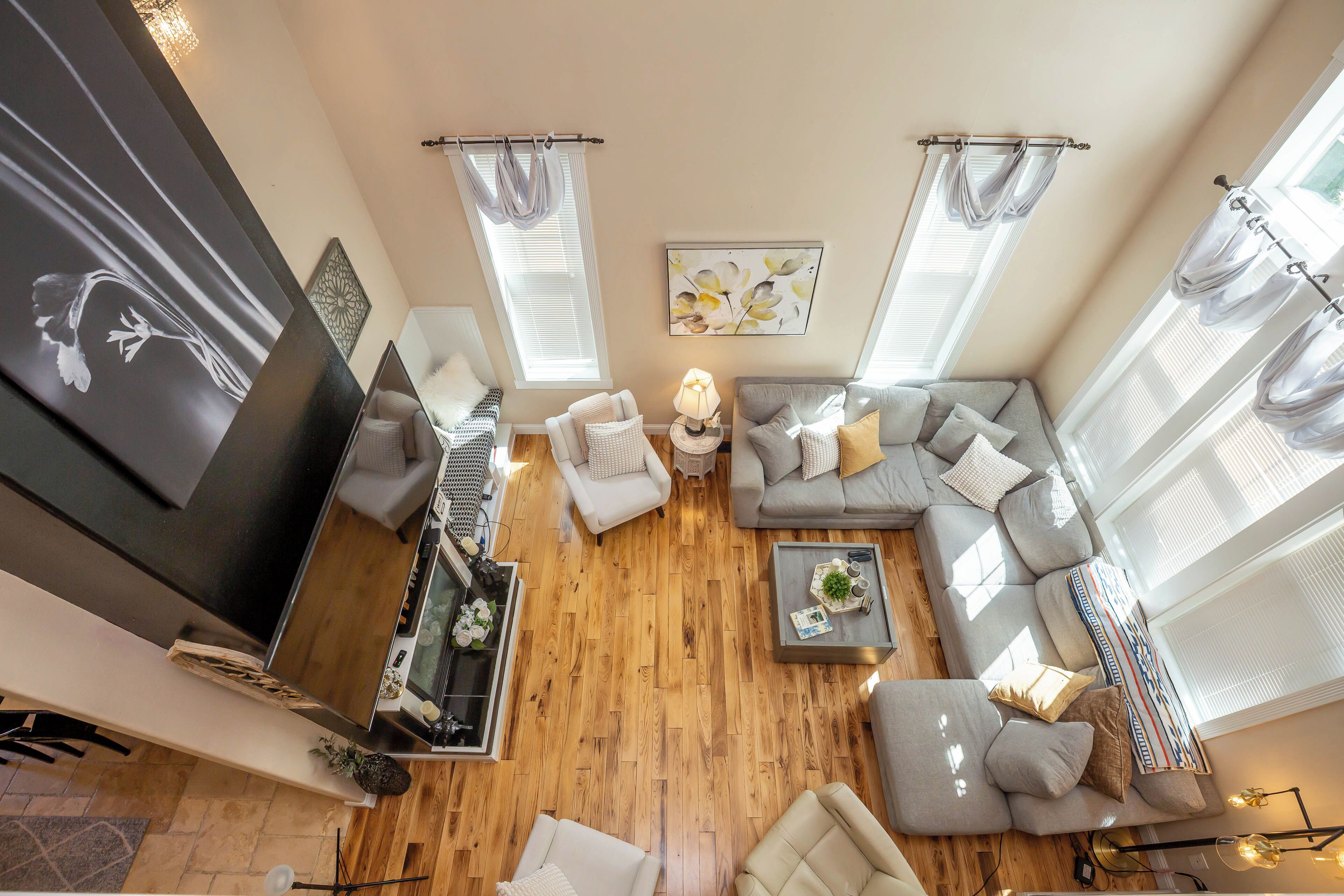 A modern living room with a sectional sofa, a coffee table, and a TV. There are two windows with curtains, a painting on the wall, and a wooden floor.