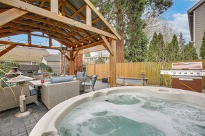 A covered outdoor patio with a hot tub, wicker furniture, and a barbecue grill. - Heated Pool, Hot Tub & Fire Pit in Ruby Park (Eugene)