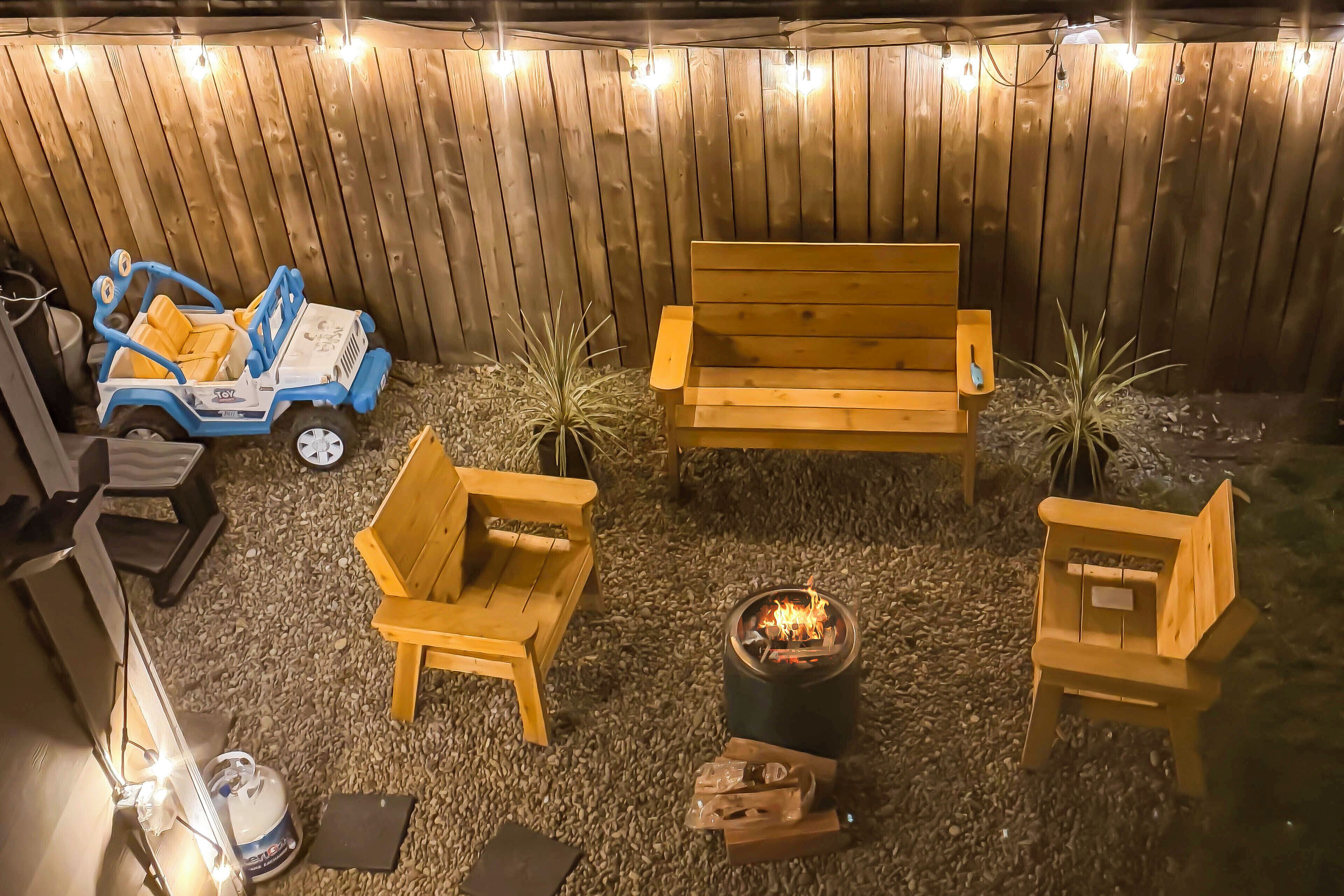 A backyard with a wooden fence, a blue toy car, a wooden bench, two chairs, a small fire pit, and string lights.