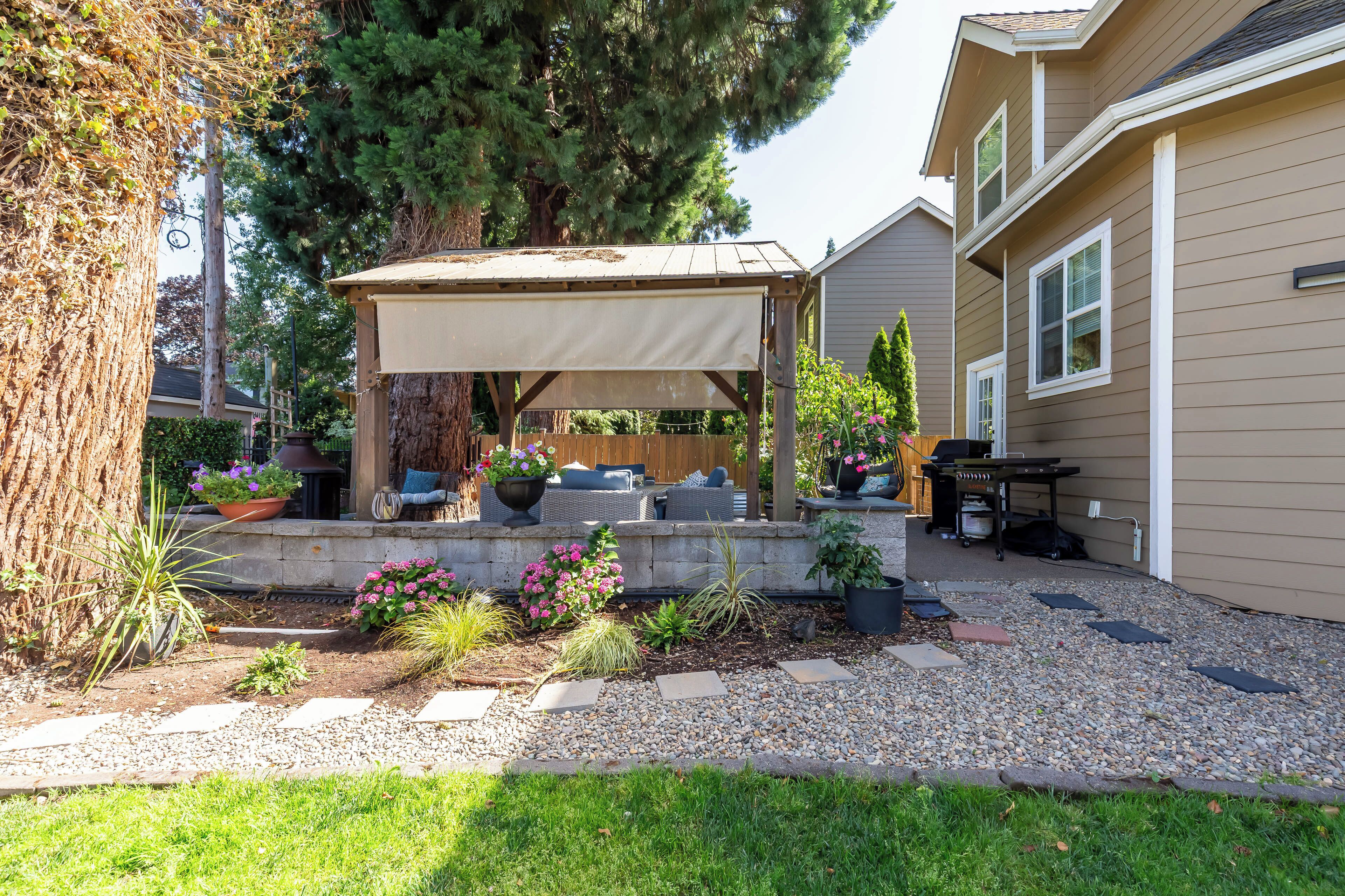 A backyard with a covered patio, a grill, and various potted plants.