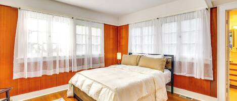 Un dormitorio con pared de paneles de madera, una cama con colcha color beige, dos mesitas de noche y un tocador.