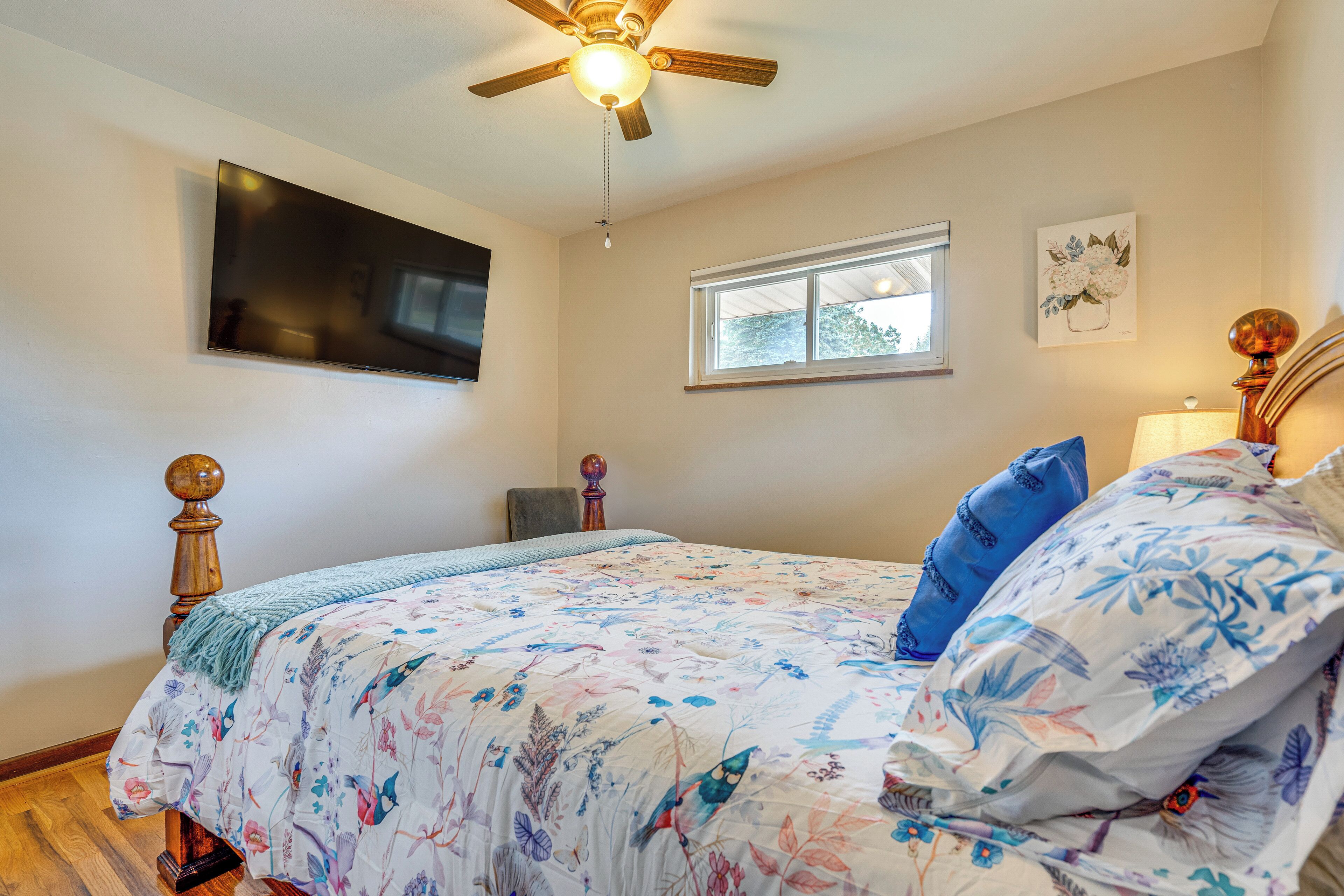 A bedroom with a bed, a ceiling fan, a TV mounted on the wall, a window, and a painting.