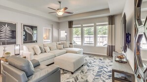 A modern living room with a sectional sofa, a ceiling fan, large windows, and a decorative rug. - Walk to Brazos County Expo: Elegant Home w/ Grill! (Bryan)
