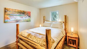 A bedroom with a wooden bed frame, a nightstand with a lamp, a window with a view of trees, and a painting of a waterfall on the wall. - Charming Donnelly Home - 1 Mi to Lake Cascade! (Donnelly)