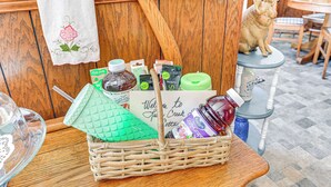 Una cesta de mimbre con varios objetos, incluyendo un recipiente verde, una botella y una tarjeta con el texto 'Welcome to Quay Creek Cottage'.
