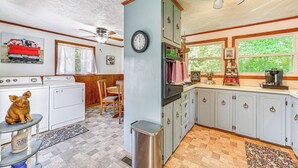Una cocina con un reloj, una estufa, un lavamanos y una zona de comedor con una silla y una mesa.