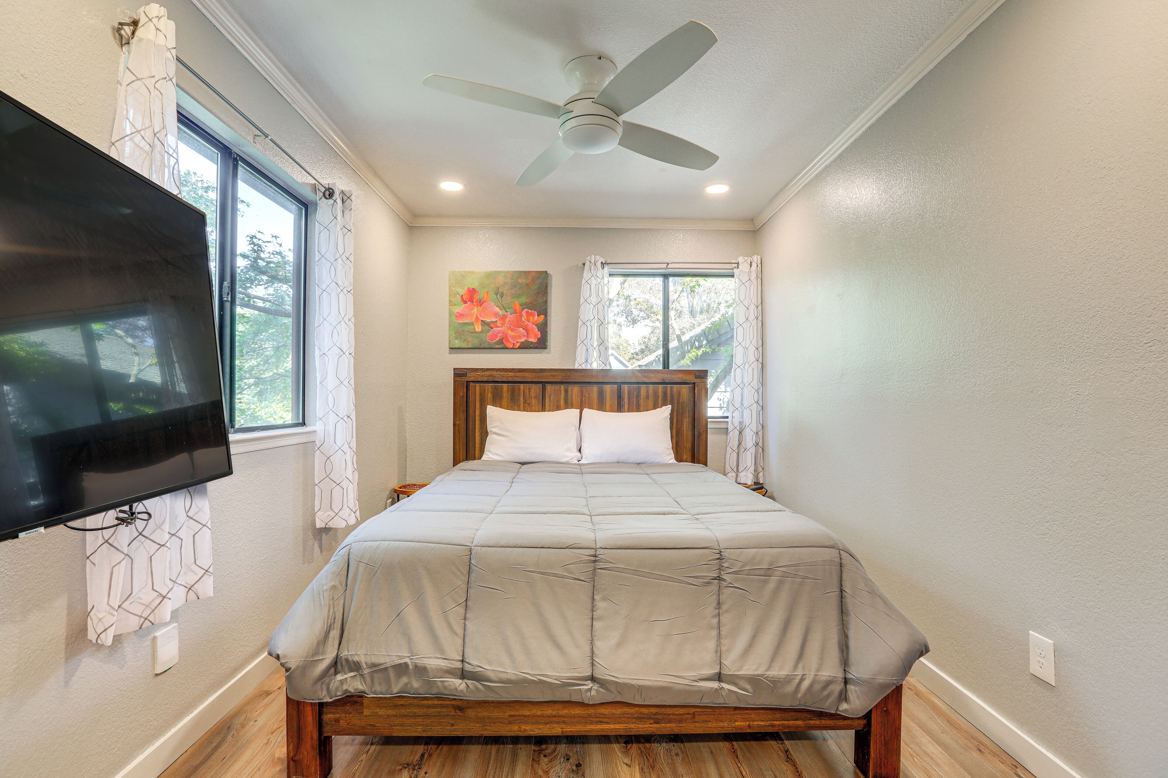A bedroom with a bed, a ceiling fan, a TV mounted on the wall, and a window with curtains.