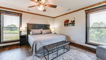 A bedroom with a bed, bench, two nightstands, a ceiling fan, and large windows with blinds.