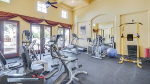 Un gimnasio amplio con diversos equipos de ejercicio, ventanas grandes y un ventilador de techo.