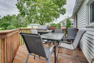 Una terraza de madera con una mesa, sillas, una parrilla y una casa al fondo.