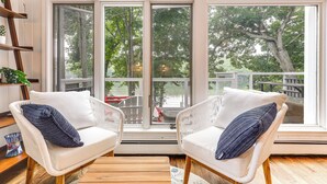 Ein gemütliches Zimmer mit zwei weißen Korbstühlen, einem Holztisch und einem großen Fenster mit Blick auf eine Terrasse und Bäume.
