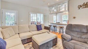 A living room with a sectional sofa, ottoman, and a gray armchair. There is a ceiling fan, a bar area with stools, and a large window with a view of greenery. - Family-friendly Harrells Home w/ Fire Pit + Kayaks (Harrells)