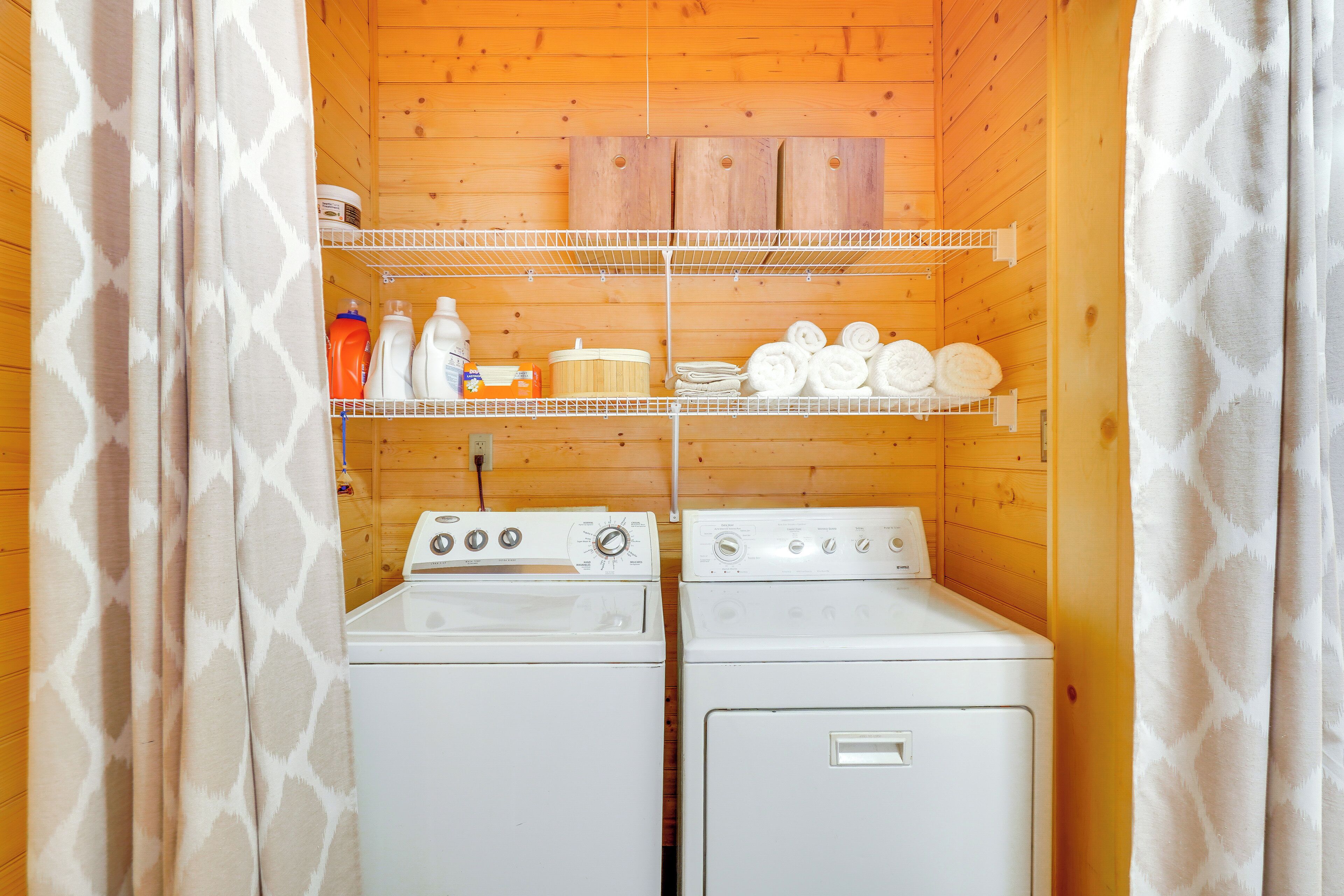 A laundry room with two washing machines, a shelf with cleaning supplies, and a wooden ceiling.