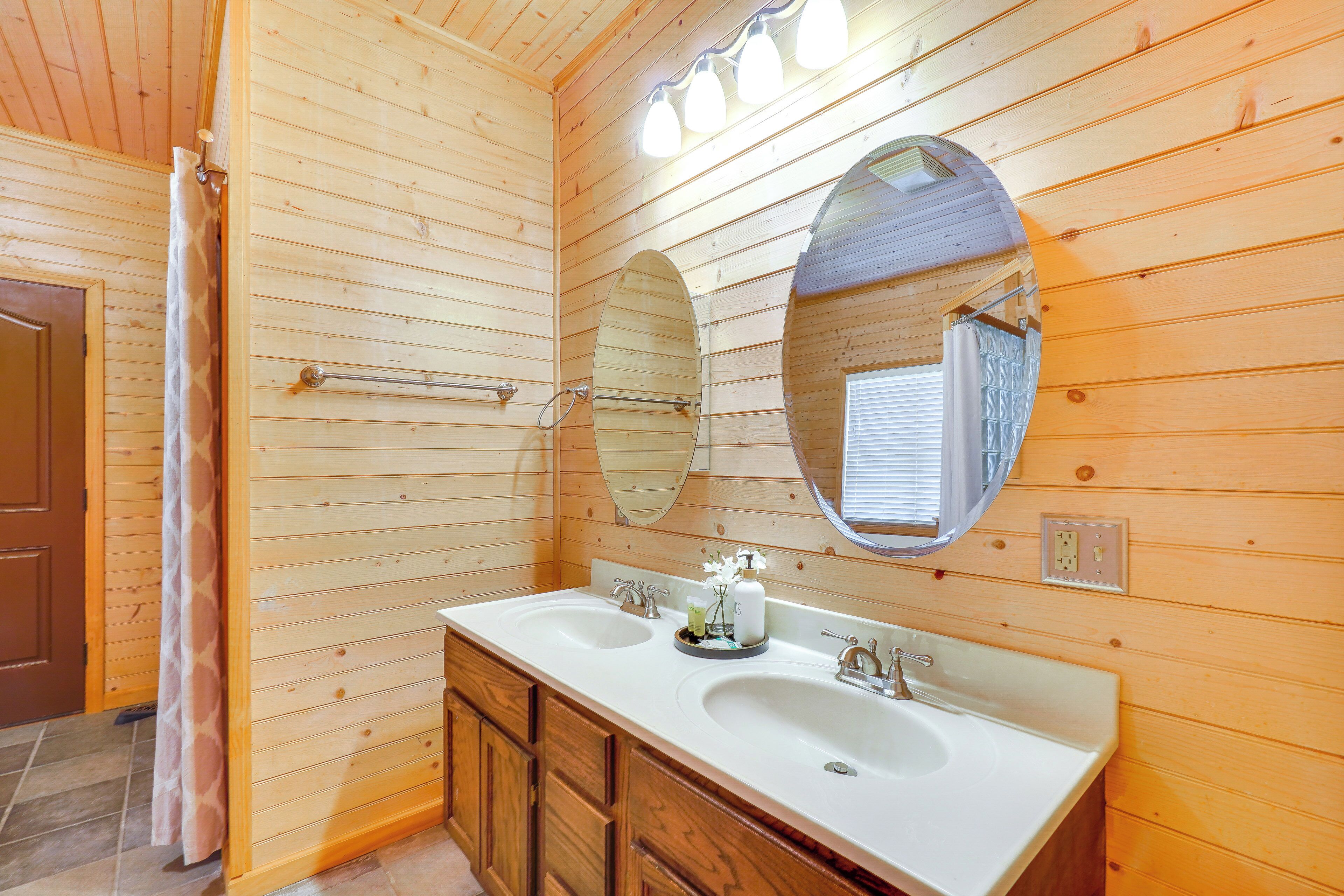 Baño de cabaña de madera con lavamanos doble, armarios de madera, un espejo grande y cortina de ducha.