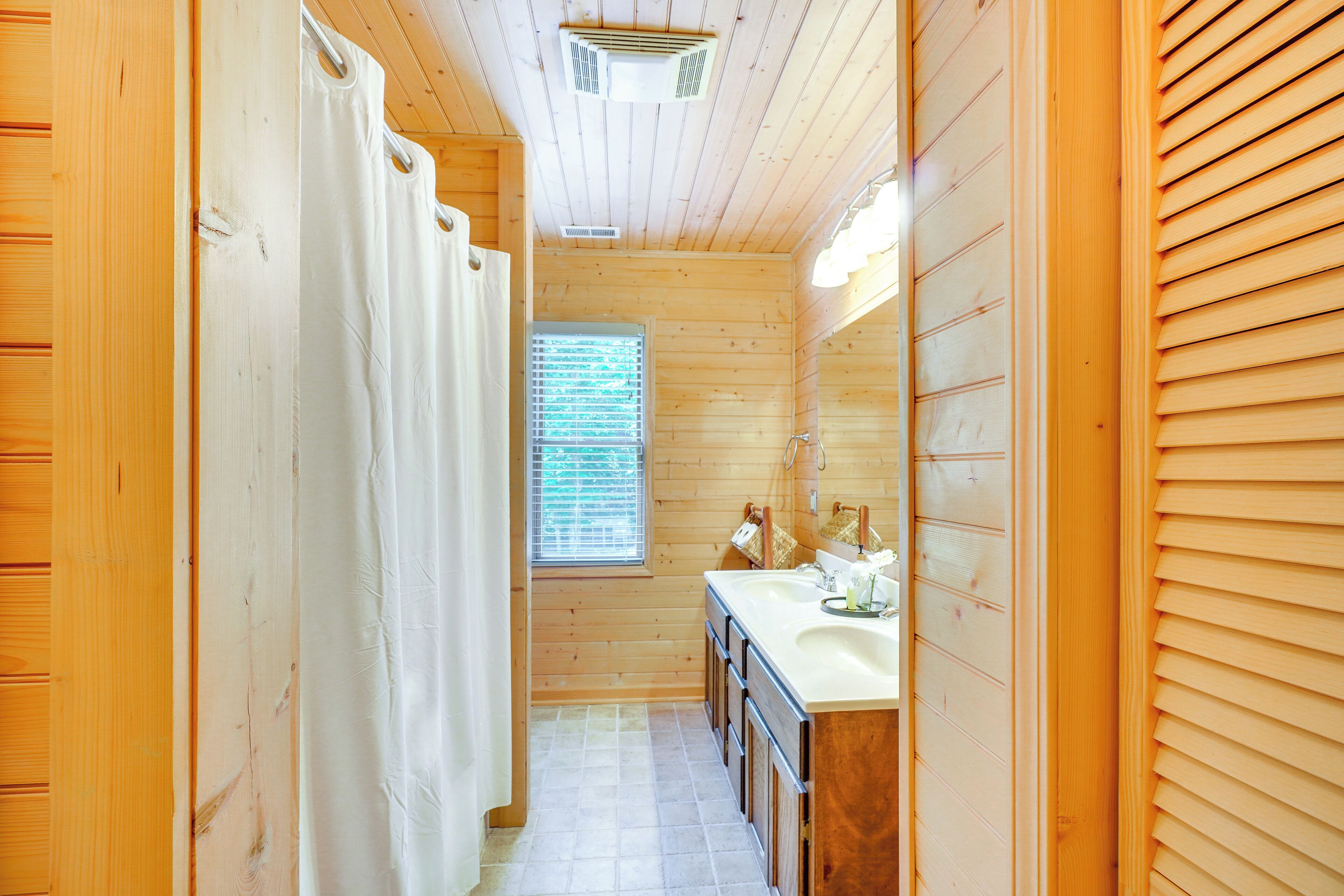 Une salle de bain rustique, avec des murs en bois, comprenant une douche avec rideau, une fenêtre avec stores, un lavabo et un miroir.