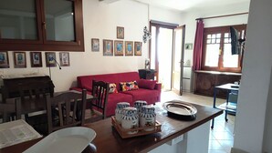 A living room with a red sofa, wooden dining table, and chairs. There are framed pictures on the wall, a kitchen area with a refrigerator, and a window with curtains.