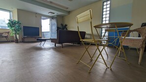 A spacious room with a large window, a blackboard, a brown sofa, a yellow folding chair, and a wicker chair. - Ace Inn Shinjuku - Hostel (Tokyo)