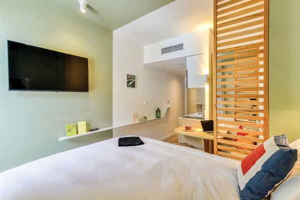 A modern hotel room with a bed, a TV mounted on the wall, a small desk with a laptop, and a wooden partition.