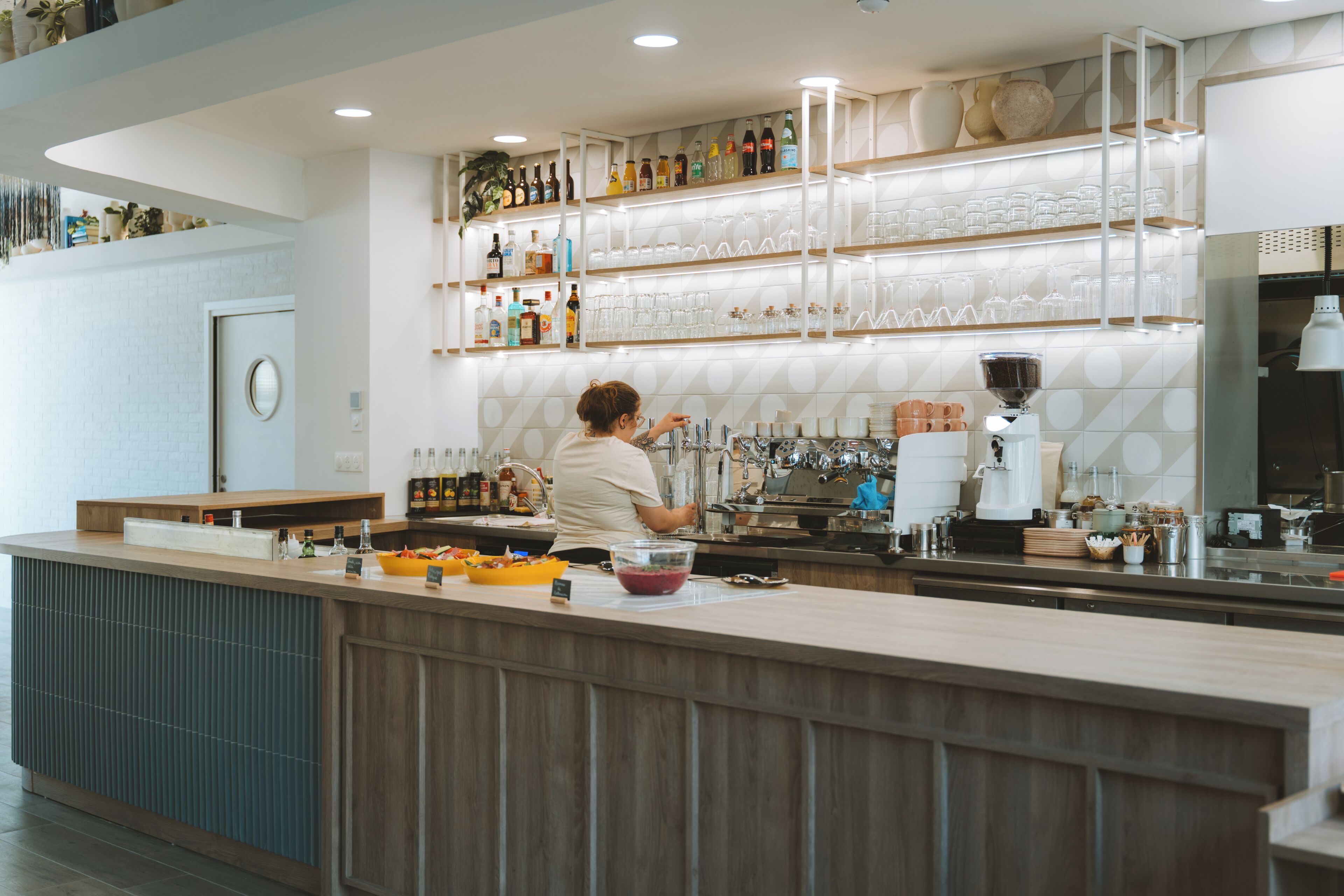 Un espace bar moderne avec un barman préparant des boissons, des étagères remplies de bouteilles et de verres, et un design épuré et contemporain.