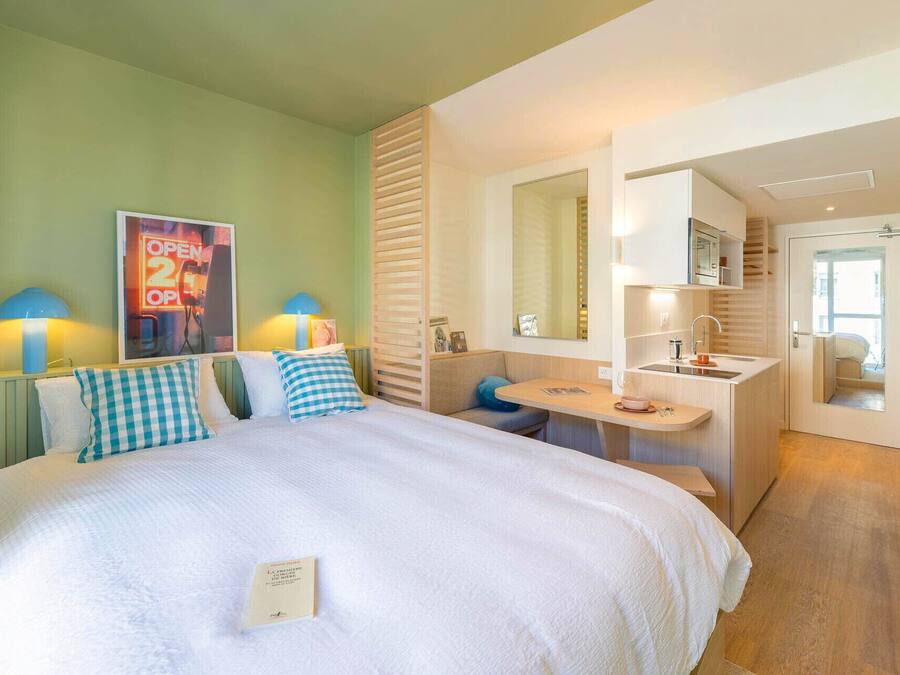 A hotel room with a bed, a desk, a mirror, and a small kitchenette.