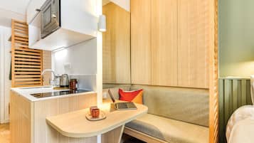 Una kitchenette compacta con microondas, lavabo y tetera, un armario de madera y un pequeño rincón de descanso con una computadora portátil y dos tazas.