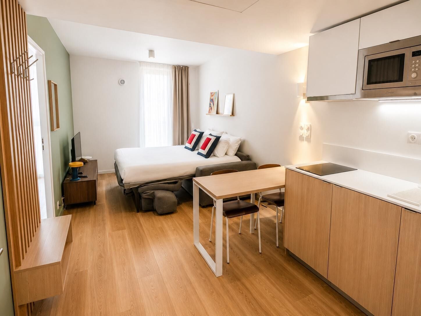 Een compacte hotelkamer met een bed, een klein bureau, een kitchenette en een raam met gordijnen.