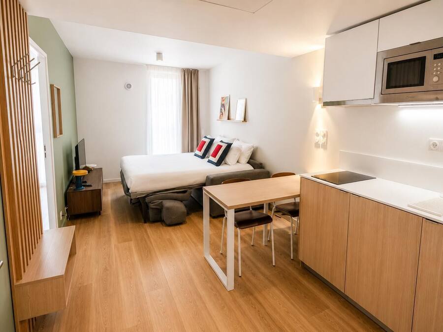Habitación de hotel pequeña y funcional con cama, escritorio, kitchenette y ventana con cortinas.