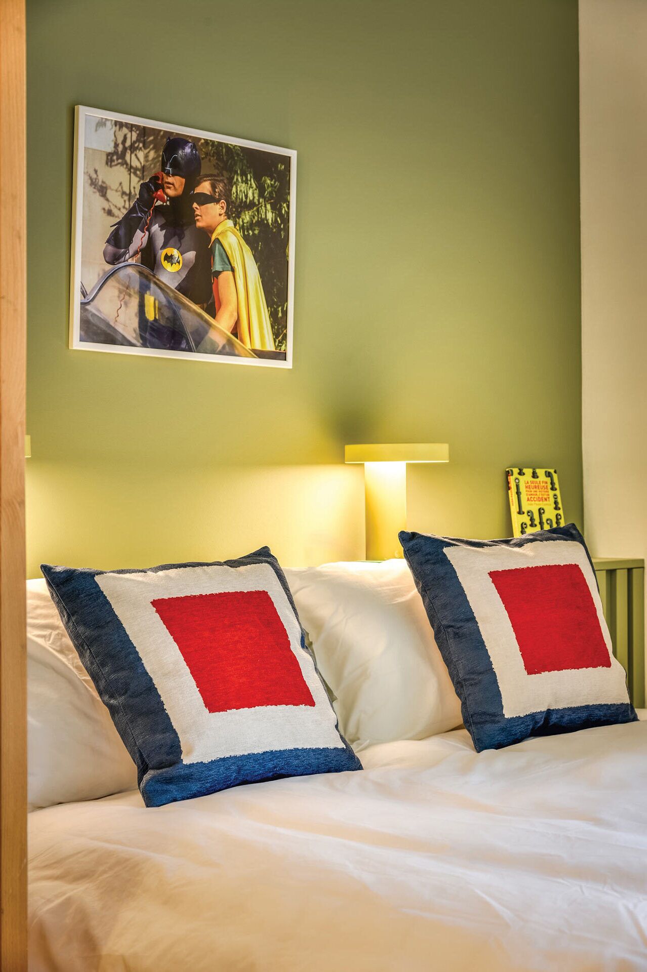 Een hotelkamer met een bed, twee kussens met een rood, wit en blauw ontwerp, een ingelijst foto aan de muur en een bedlamp.