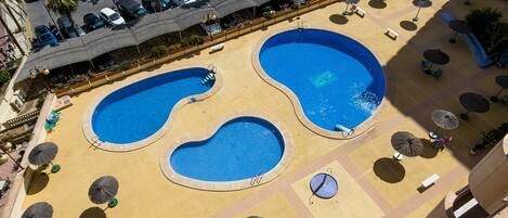 A rooftop pool area with two large pools, sun loungers, and umbrellas.
