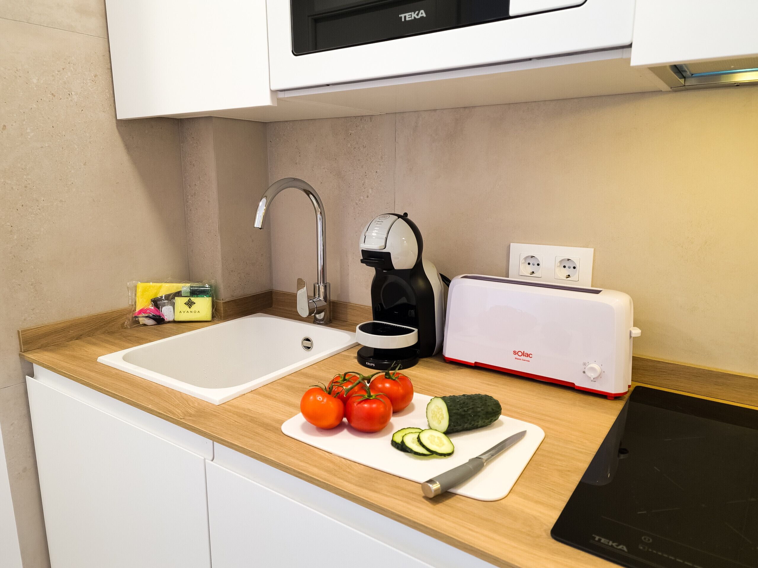 A kitchen with a sink, a toaster, and a kettle. There are tomatoes and cucumbers on the counter.