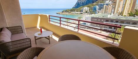 A balcony with wicker furniture, a table, and a bottle of wine. It overlooks a beach with a rocky mountain in the background and several buildings.