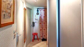 Un couloir étroit avec un tabouret rouge, une porte dotée d’un luminaire et un mur orné d’un tableau encadré.